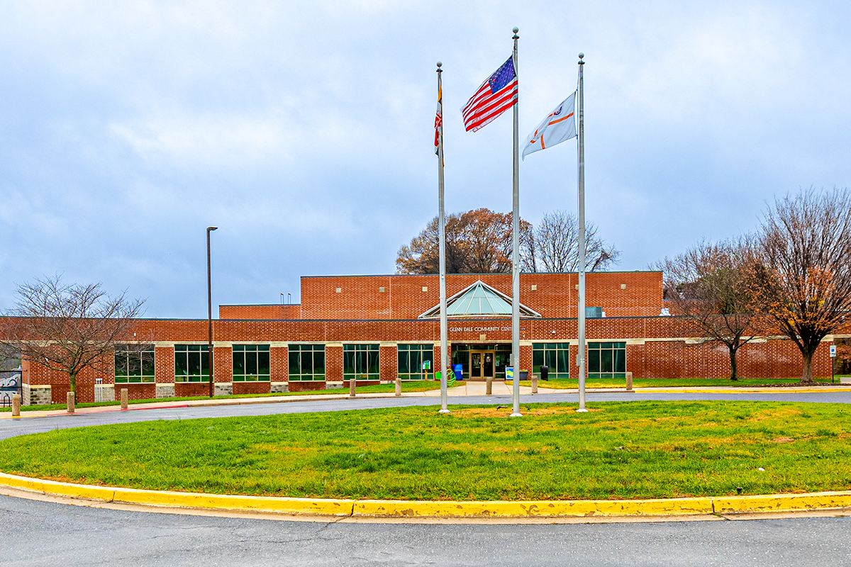 Exterior view of the circular driveway in front of a large, long, red brick building. Inside the circular drive , three flags fly on flagpoles in the grass, including the U.S. flag and the Maryland state flag. A sidewalk leads to double glass entrance doors in the building. Silver lettering over the entrance reads GLENN DALE COMMUNITY CENTER.