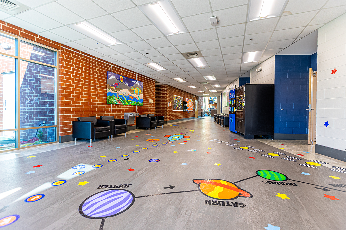 View from a lobby area down a long hallway with one red brick wall and one cinderblock wall painted white and blue. The floor has an elaborate design of planets, alphabet letters, stars, footprints, and a rocket ship floor decals. There are two vending machines against the cinderblock wall, and black armchairs along both walls.