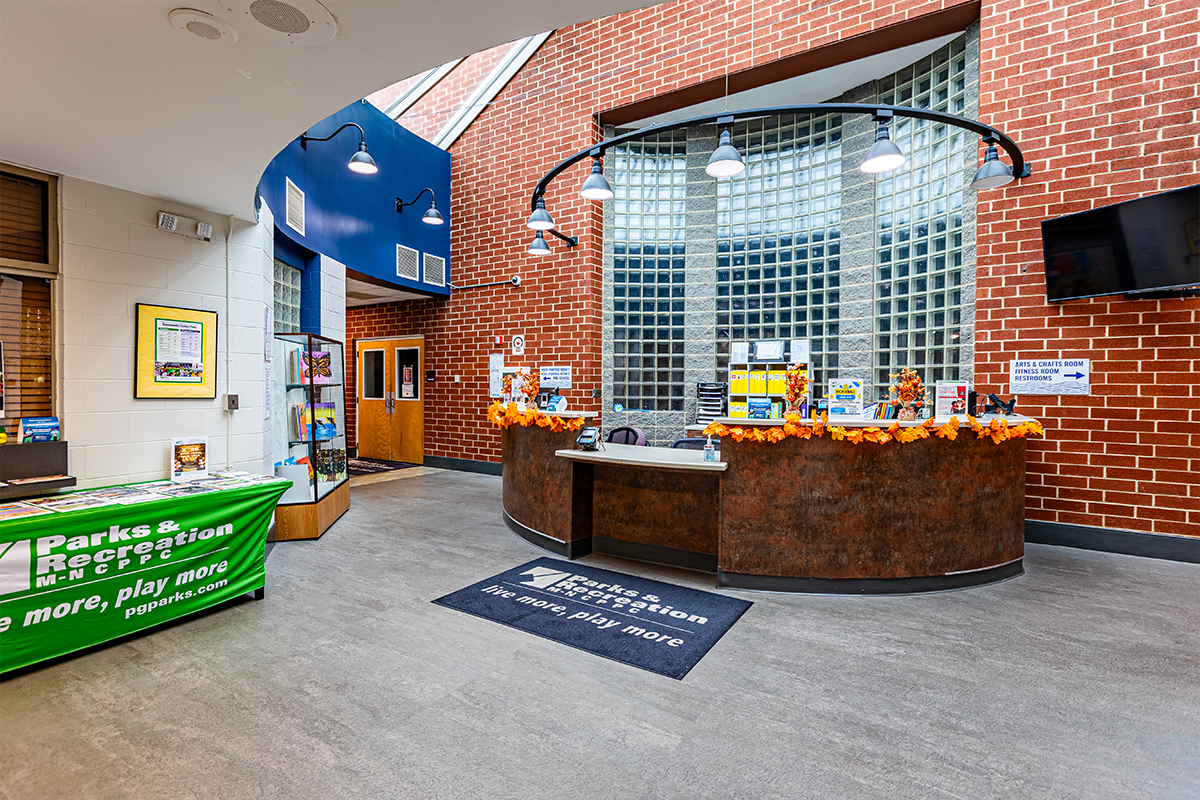 A lobby space with a round, brown reception desk situated in front of a curved, glass block window in a red brick wall. To the left is an information table with a green Parks and Rec tablecloth covered with flyers. A sign on the wall has an arrow and the words Arts & Crafts Room, Fitness Room, Restrooms. The reception desk is decorated with autumn leaf decorations.