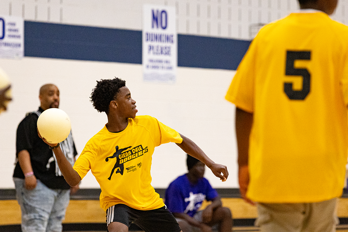 In a gymnasium, a young man in a yellow jersey leans back with a white ball in his right hand, preparing to throw it. The jersey reads Glenn Dale Dodgeball. Another person in a yellow jersey stands near him. In the background are several spectators on wooden bleachers under a sign mounted on the wall that says NO DUNKING PLEASE.