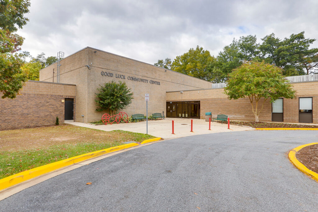 Exterior view of the entrance to Good Luck Community Center, a large brick building with benches and a bike rack out front.