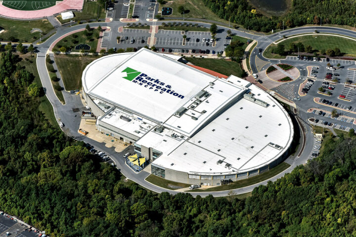 Aerial view of a large oval-shaped building with a white roof, emblazoned with a giant Parks and Recreation logo. There are several parking lots next to it on one side, and a wooded area on the other side.