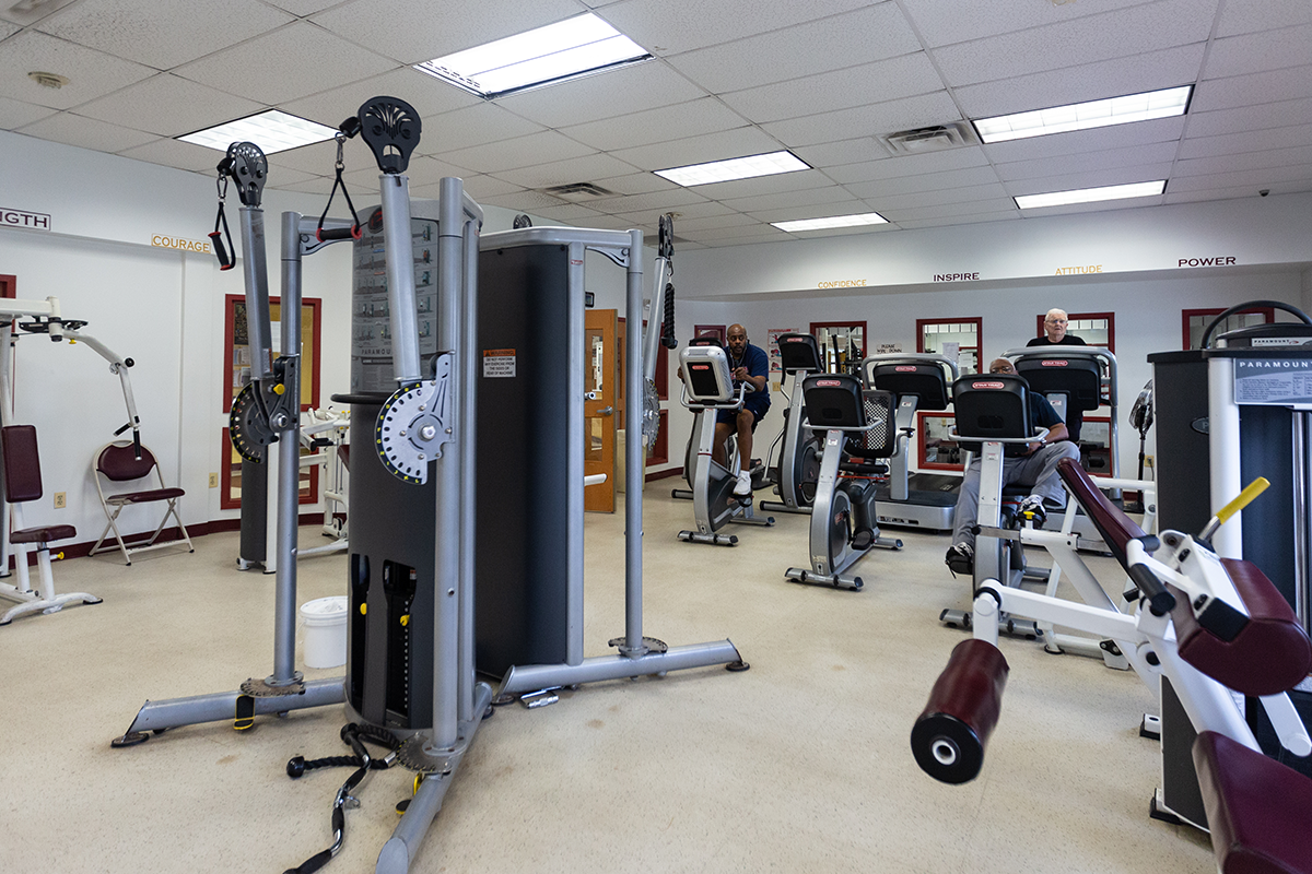 A fitness room with many pieces of exercise equipment, including stationary bikes, treadmills, and weight machines. Encouraging words are written along the top of the gray walls, including INSPIRE, COURAGE, POWER, and STRENGTH.