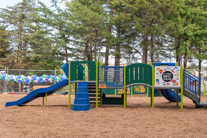 Side view of a long piece of play equipment at an outdoor playground, with slides at both ends, multiple sets of stairs, platforms, spinning features, and a curved bridge. In the background is a chain-link fence decorated with balloons.