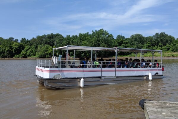 A white pontoon tour boat in a brown body of water with trees along the far shore. The boat has a shade canopy and rows of benches which are all filled with people.