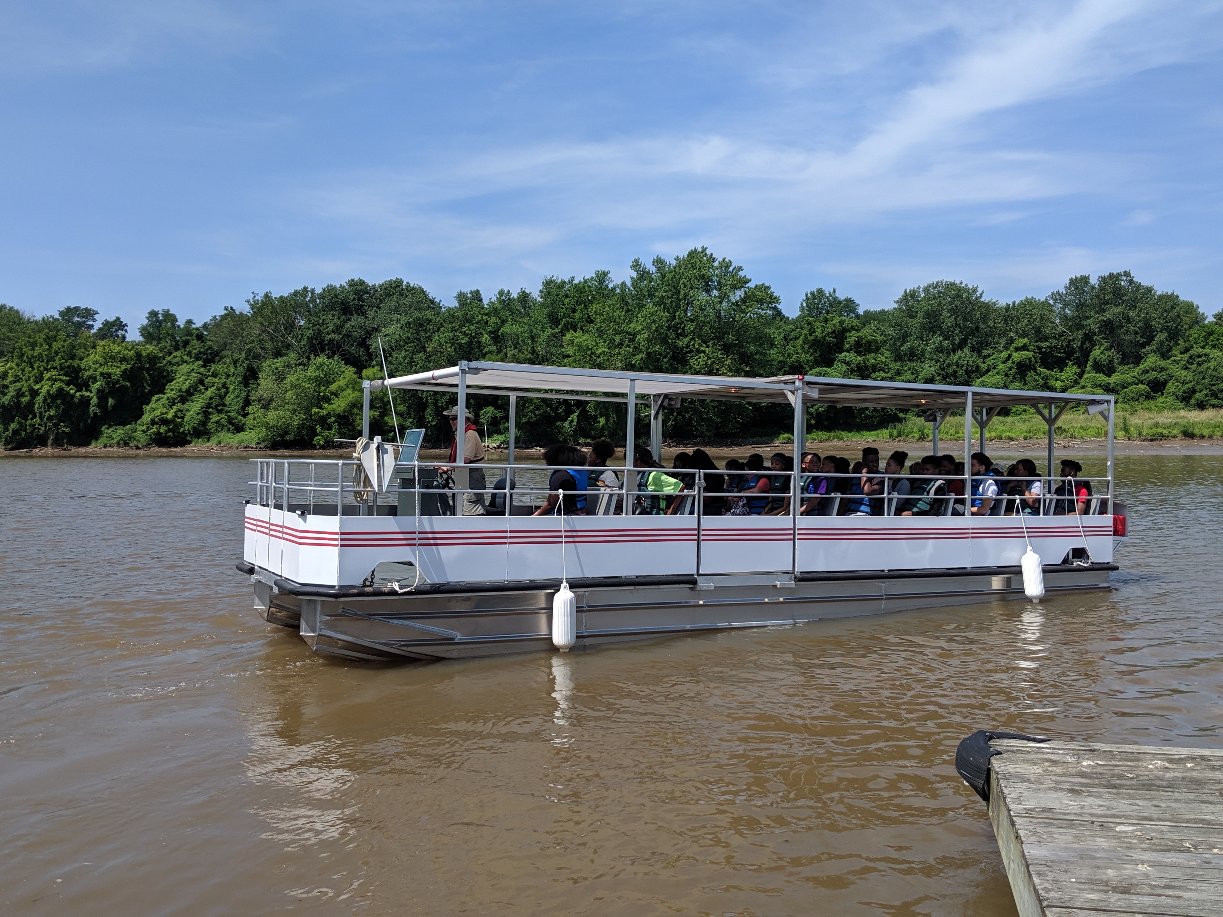A white pontoon tour boat in a brown body of water with trees along the far shore. The boat has a shade canopy and rows of benches which are all filled with people.