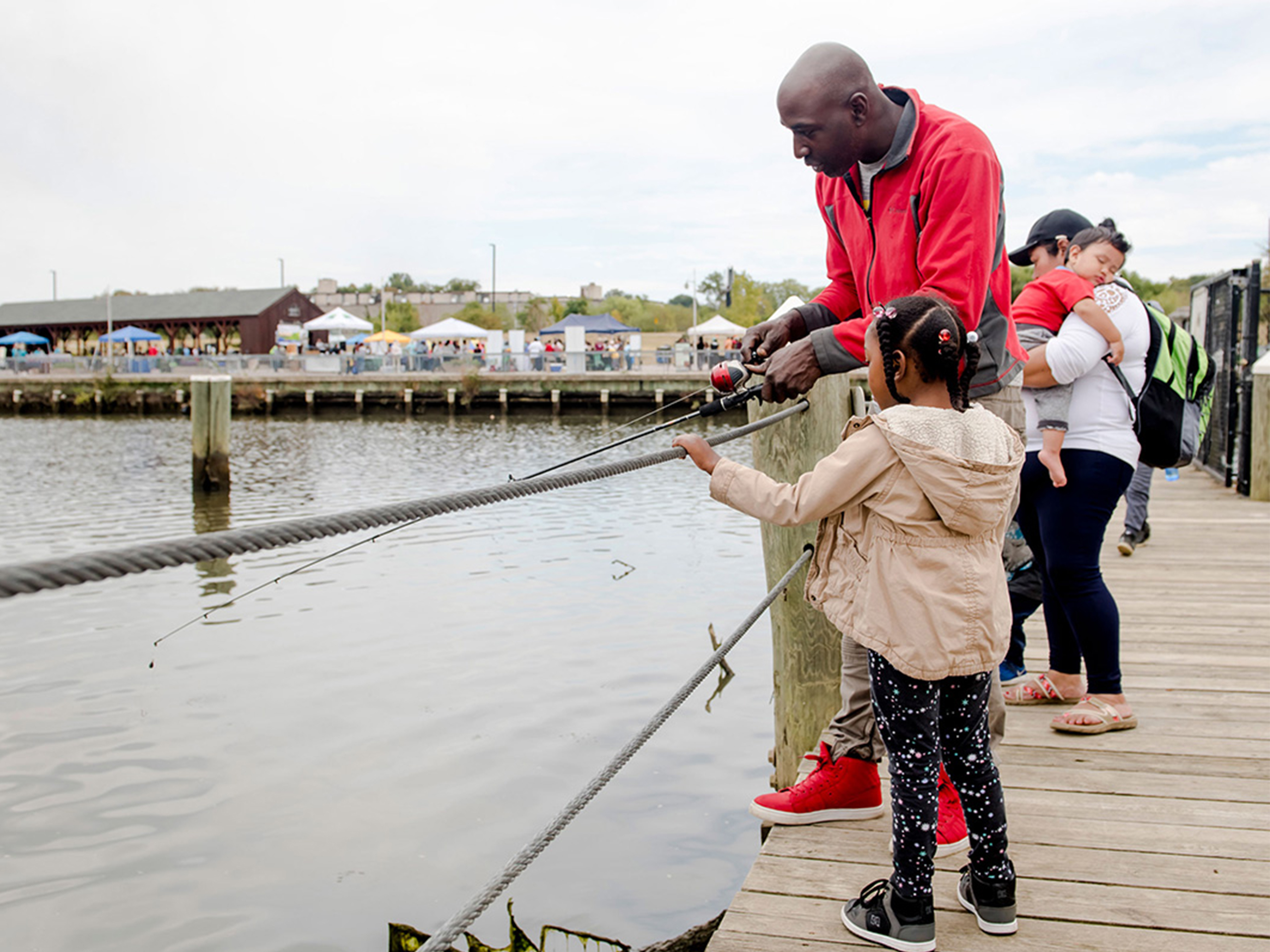 A man holding a fishing pole stands on a wooden pier next to a child. In the distance, a row of event tents is set up between a picnic shelter and the water's edge.