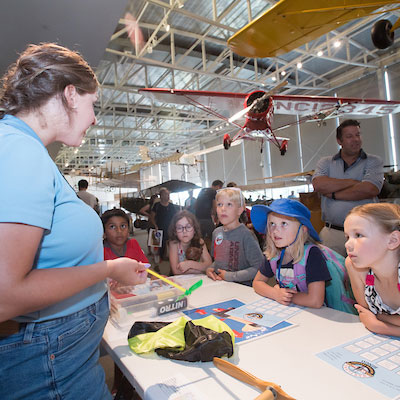 Five children with worksheets in front of them stand in front of a table, where a staff member is holding a stick with a small propeller attached. Behind them in a large hangar space, several vintage aircraft are suspended from the ceiling.