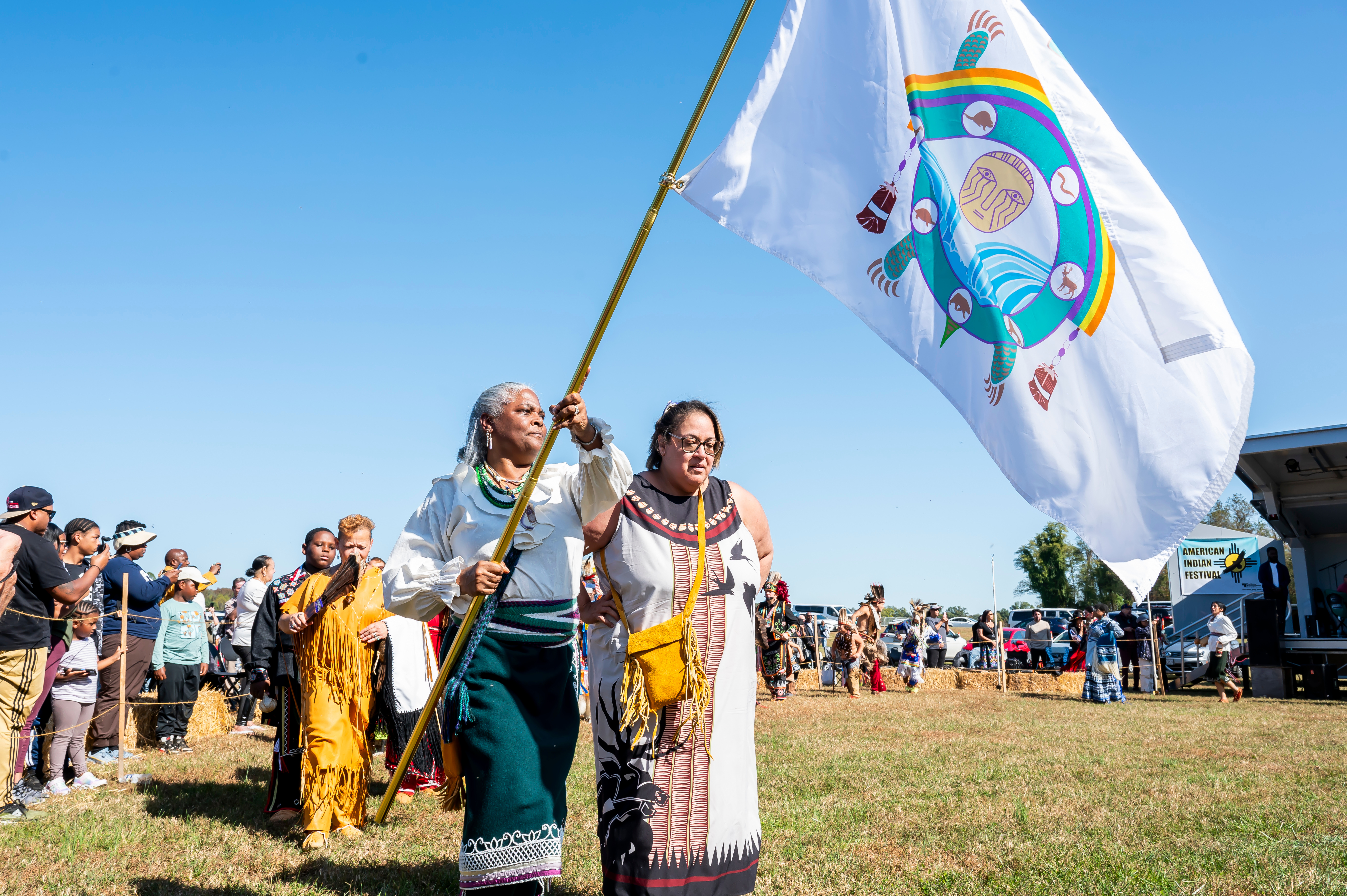 In a grassy field, two women in traditional Native American dresses lead a procession. One of them carries a large flag on a pole.