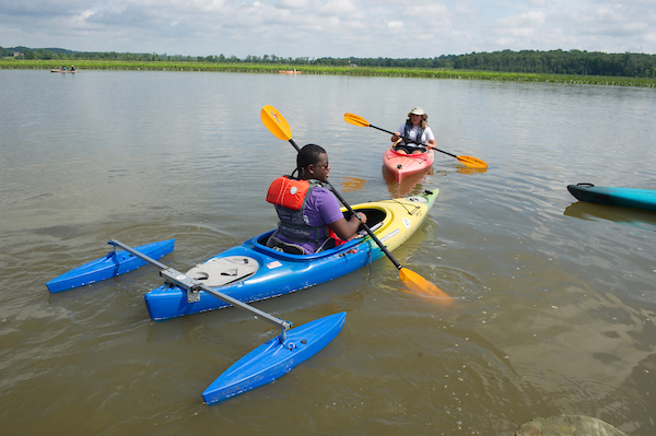 In a large body of water, a man paddles a kayak that has two stabilizing pontoons attached to the back end.
