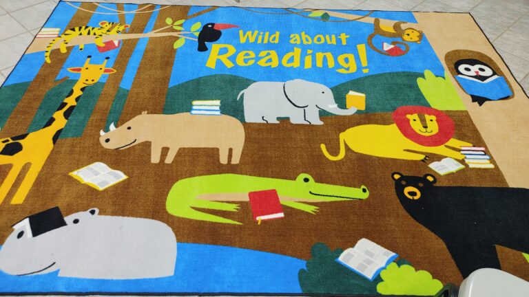 A colorful rug on a beige, tiled floor, with cartoonish images of wild animals in a nature setting with stacks of books, and the words WILD ABOUT READING at the top.