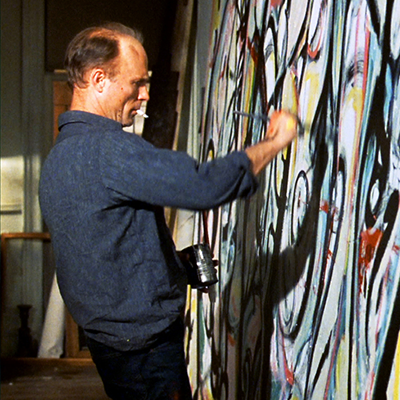 Publicity still from the 2000 film POLLOCK. A man (played by actor Ed Harris) with a cigarette in his mouth paints on a canvas the size of the wall covered in a colorful, abstract design.