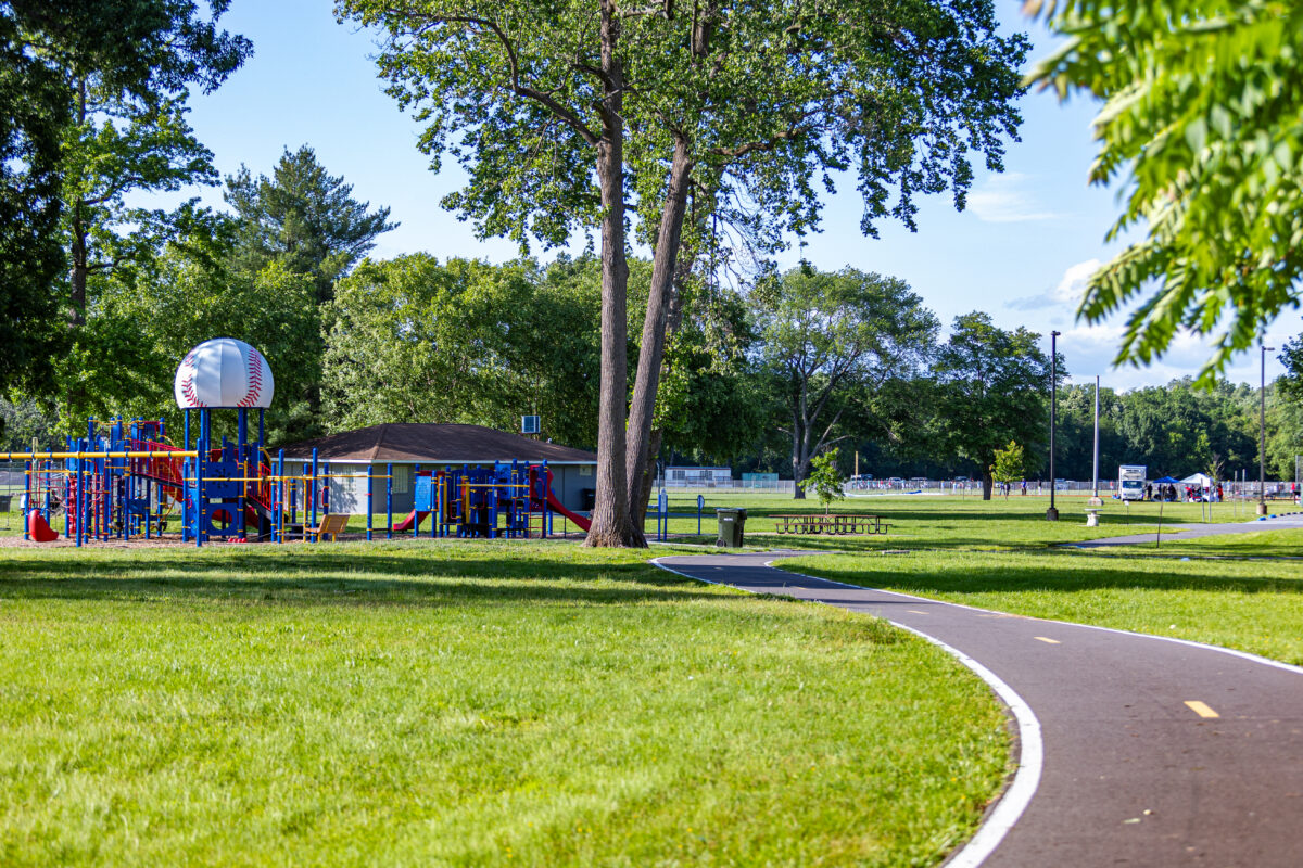 A paved asphalt trail with a dotted line down the middle curves through a grassy lawn toward a red and blue outdoor playground. There are multiple large play structures with slides, platforms, monkey bars, and more. The central tower of one play structure is topped with a roof resembling a giant baseball. Near the playground are several picnic tables, and in the distance are sports fields.