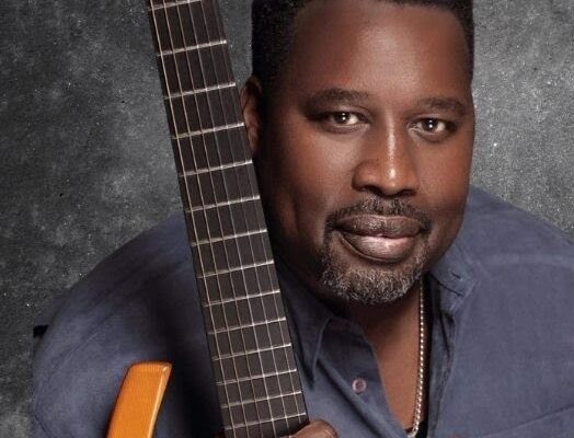 A man with a close-cropped beard, wearing a blue shirt and a silver necklace, poses with one hand around the neck of an electric guitar.