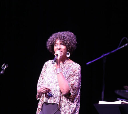 A woman with curly hair wearing a loose, patterned blouse, sings on a dark stage into a handheld microphone, lit by stage lighting.