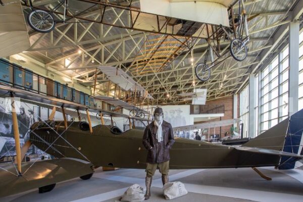 A mannequin dressed in historical pilot gear, including goggles, a brown leather cap, a brown leather jacket, and a white silk scarf, posed in front of a dark green, two-man propeller plane in a large museum gallery. Three more historical aircraft are displayed in the gallery, some suspended from the ceiling.