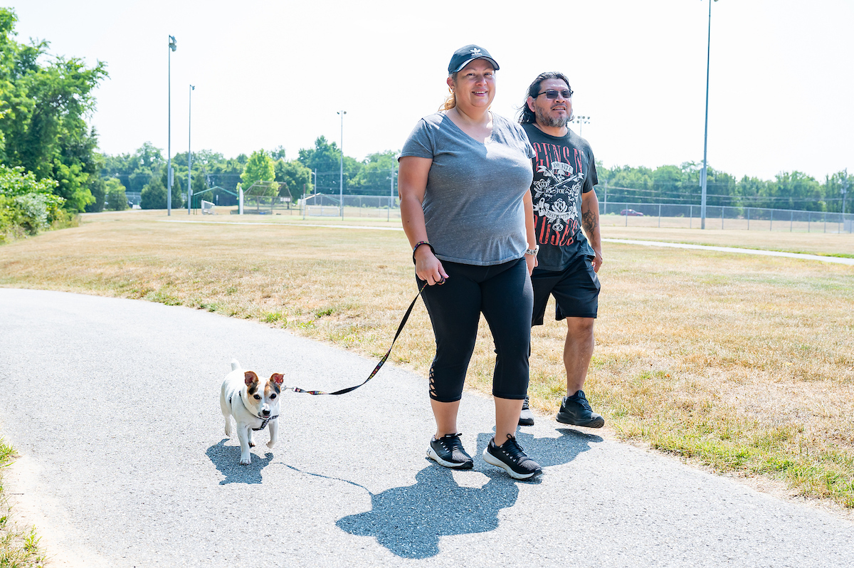 Two smiling adults walk along a paved trail with a small dog on a leash. Next to the trail is a flat, grassy lawn, with a sports field in the background surrounded by a chain-link fence.