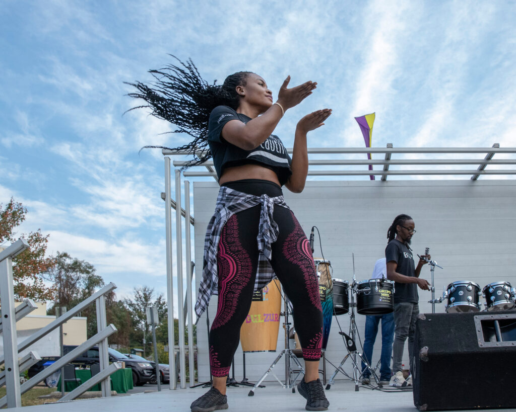 A woman with long hair dances on an outdoor stage. Behind her are multiple hand drums and a drum kit.