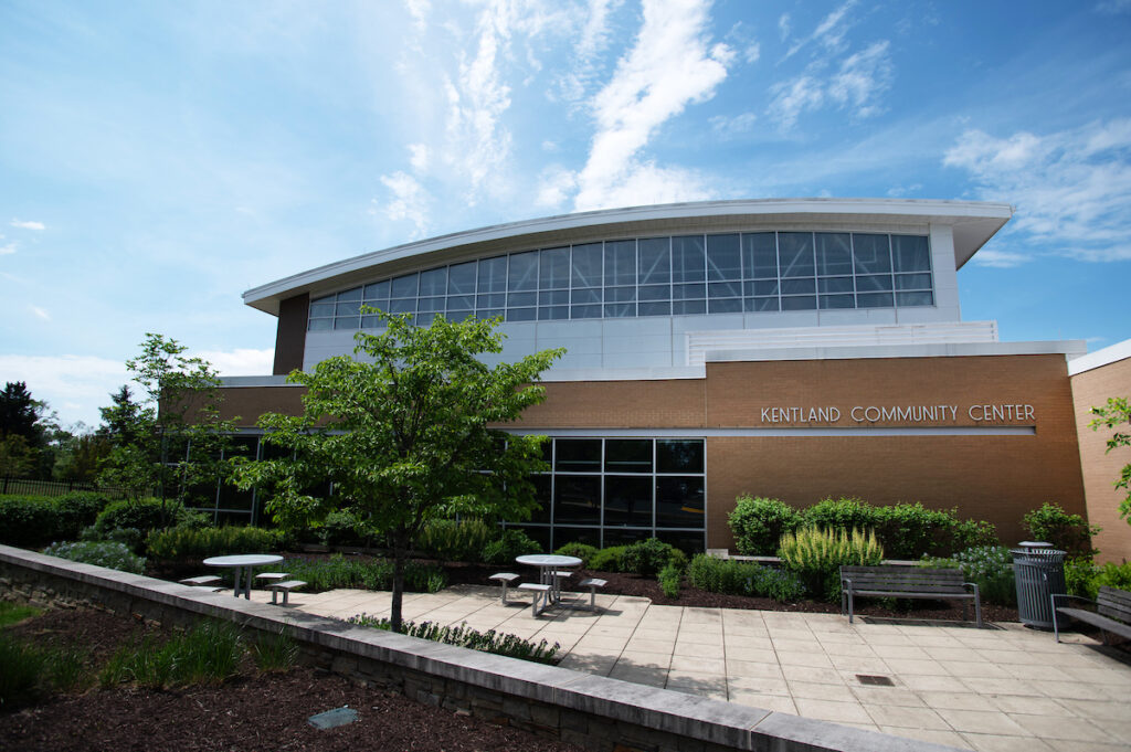 A large brick, glass, and metal building with a landscaped patio. Letters on the building read Kentland Community Center. On the patio are benches and café tables.