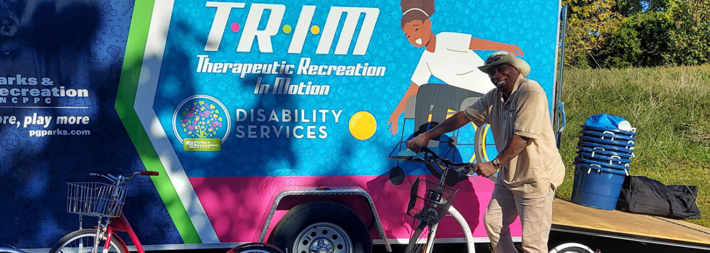A smiling staff member in a tan Parks and Rec shirt stands on a white kickbike in a parking lot. Next to him is a large truck or trailer with the words TRIM: Therapeutic Recreation in Motion on the side, and the Parks and Rec logo. A red kickbike stands on the ground nearby.
