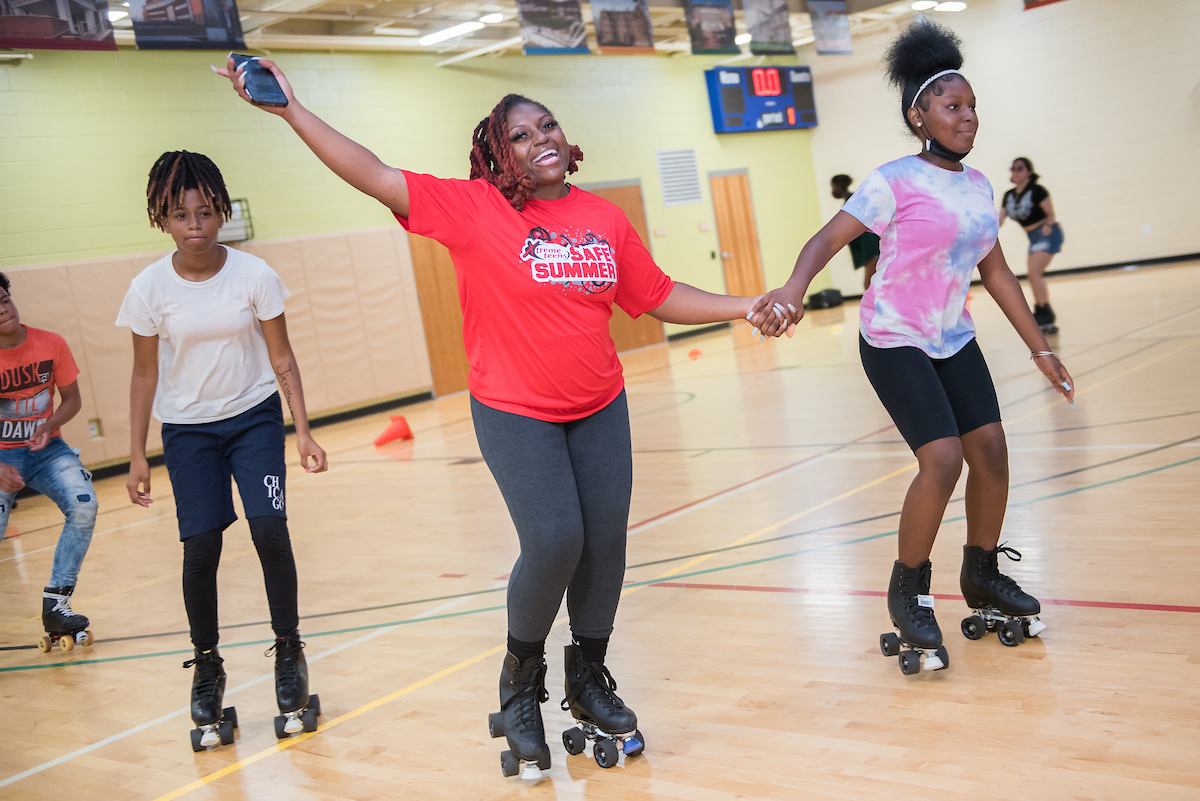 Two women hold hands while roller skating in a gymnasium. One wears a red "Safe Summer" t-shirt. Behind them other youths are roller skating.