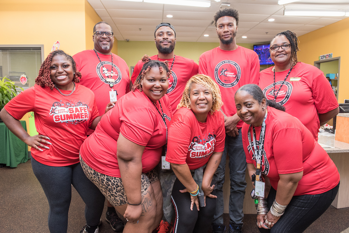 Eight smiling adults in red "Safe Summer" shirts and nametags pose together for a photo in a lobby area.