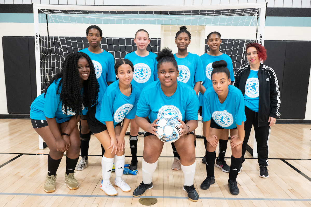Eight girls and their coach, all in matching turquoise t-shirts, pose in a gymnasium in front of a futsal goal with a white net. A girl in front holds a futsal ball in her hands.