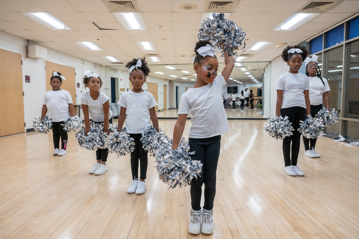 In a dance studio with hardwood floors and one mirrored wall, six girls in matching black and white outfits stand in a V-shaped formation holding silver pom-poms.