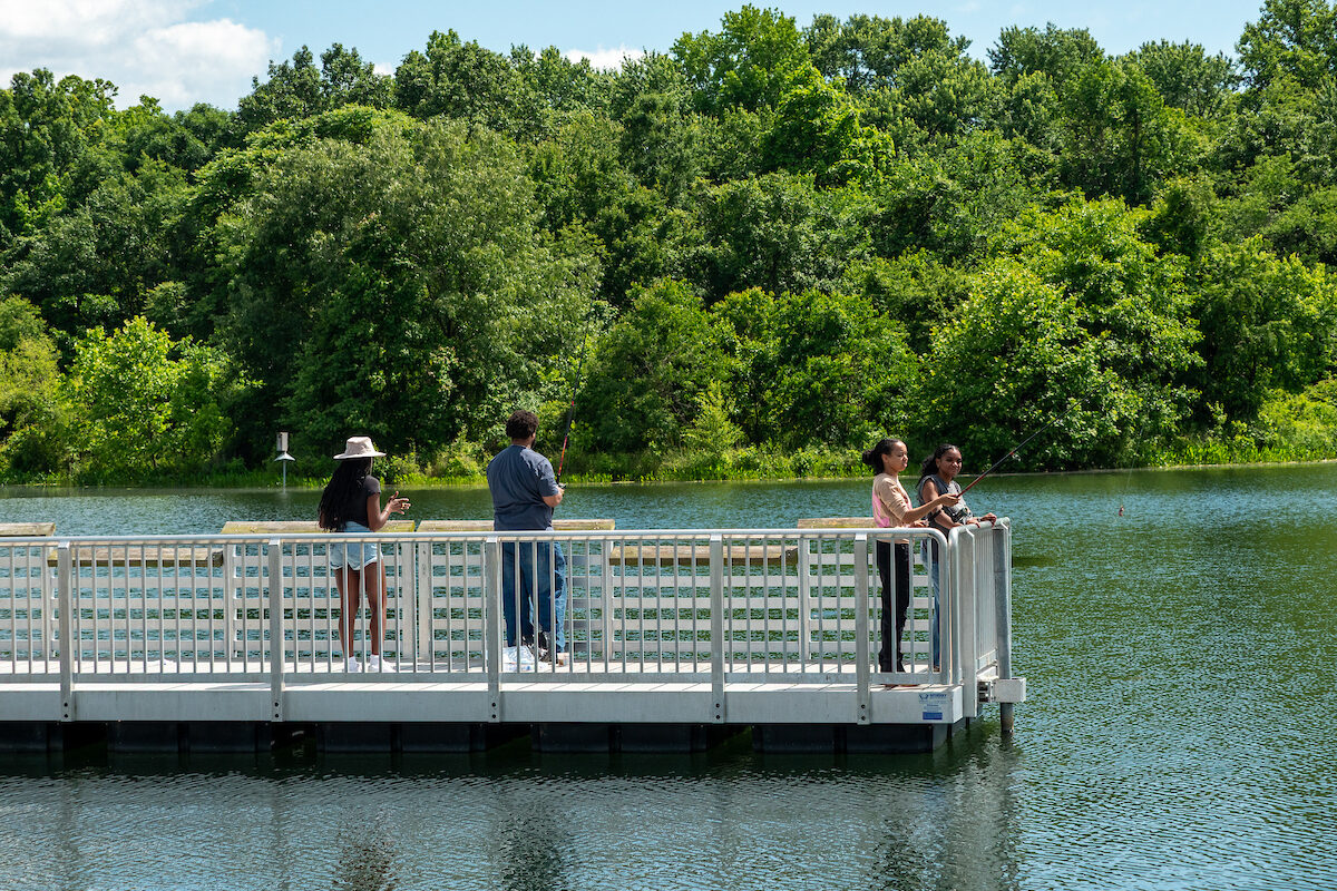 Four people fish with fishing poles off a white pier jutting out into a lake. The water is calm, with very tiny ripples from the breeze. Across the water is a forested area.