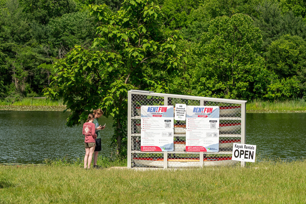 In the grass at the edge of a lake, a man and woman stand next to a kayak rental cage with a sign next to it that reads KAYAK RENTALS OPEN. The man is looking at his phone. Signs on the kayak rental cage have instructions in English and Spanish, and QR codes. The cage contains a stack of kayaks with orange tops and white bottoms.