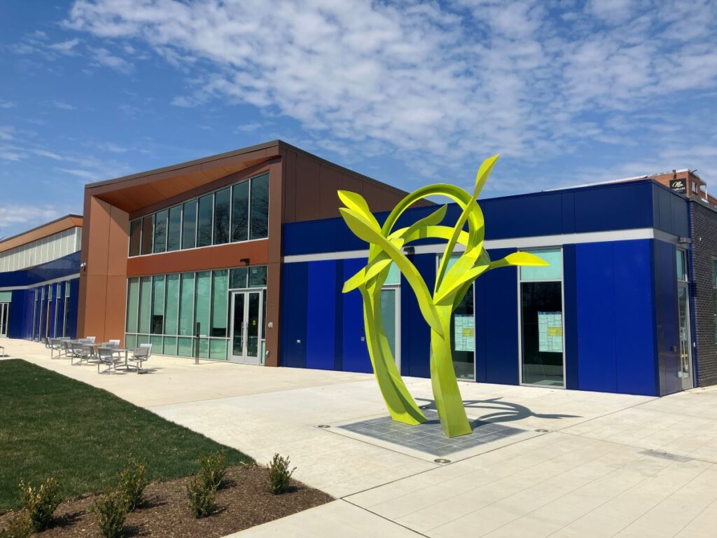 A large, modern, glass, brick, and metal building with blue panels. In front is a large, neon green sculpture that resembles plant stems and leaves.