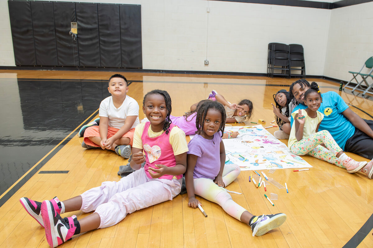 Seven kids and one camp counselor sit and lie on the floor of a gymnasium, working on a large paper banner. There are colorful markers and colored pencils scattered around. They are all posing and smiling for the camera.