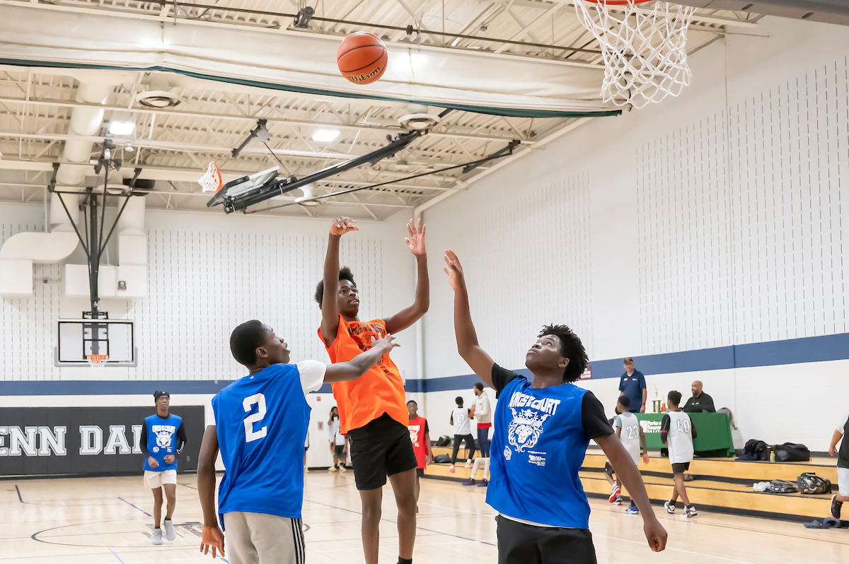 In a large gymnasium with the words GLENN DALE painted on the far wall, four teenage boys play basketball. One is in midair, having just thrown the basketball toward the hoop. Behind them to the right are about a dozen people on low, wooden bleachers/benches, and a staff member sitting at a table.