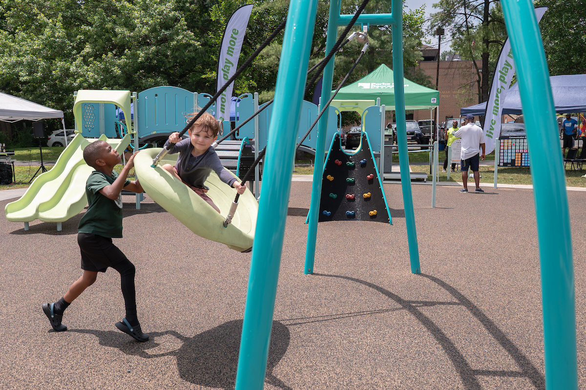 At an outdoor playground, one boy pushes another on a large, plastic, dish-style swing. Behind them is a large play structure with slides, platforms, and a climbing wall. Beyond the playground, several event tents are set up in the grass.