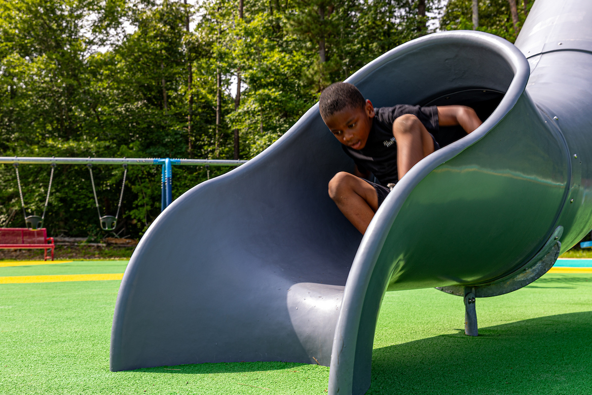 A child emerges from the bottom of a tube slide on an outdoor playground. Behind the slide, a swing set and a red bench are visible, with tall trees behind them.
