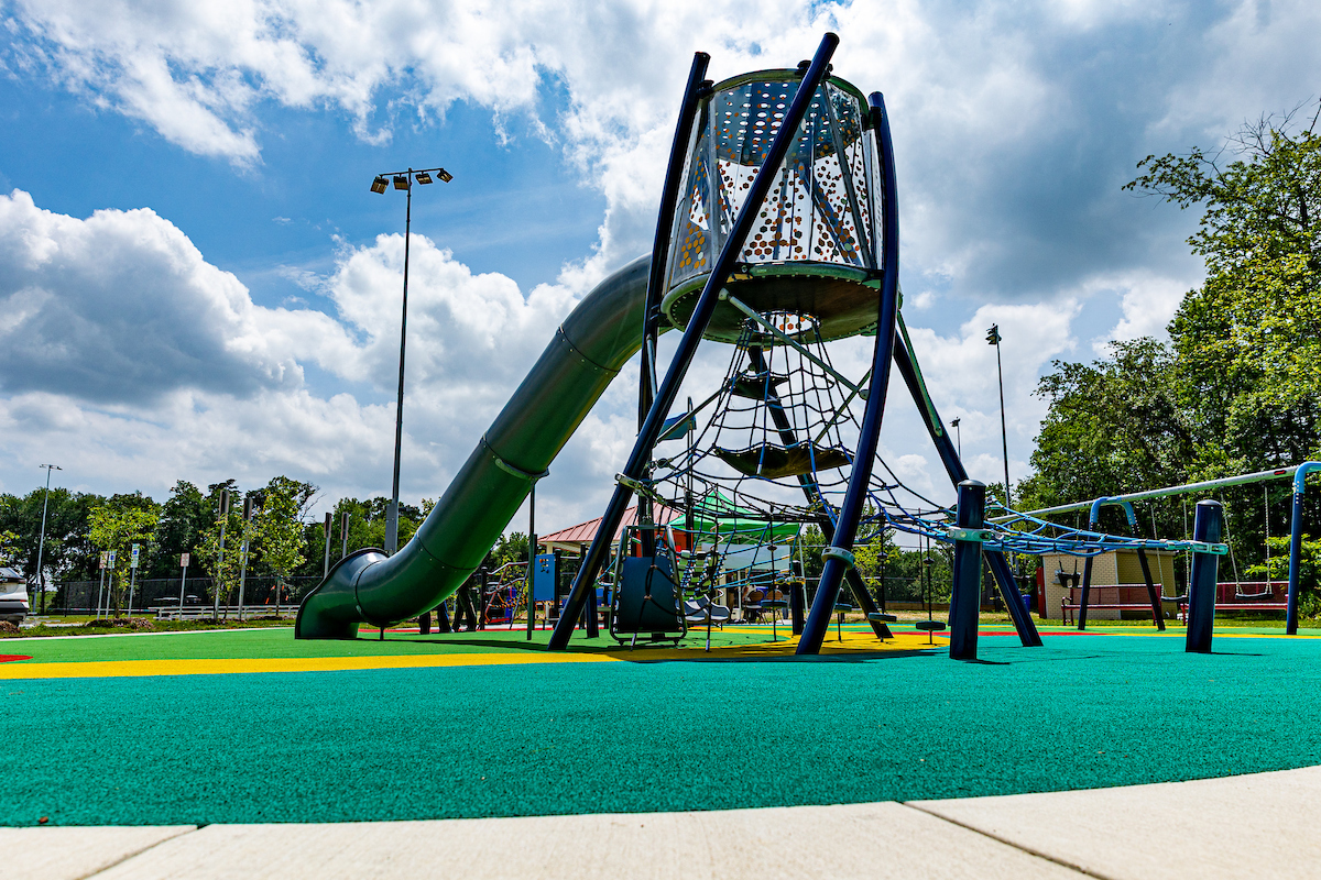 A tall play structure on an outdoor playground featuring a tube slide down from a high platform encased in plexiglass walls, and a rope web below the platform as the only visible means for climbing up.