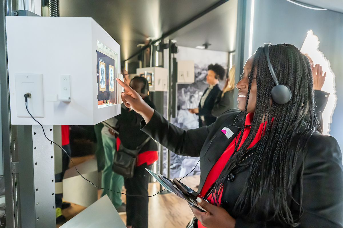 A woman smiles while selecting an image on a small video monitor, wearing headphones connected to it. There are two historical portraits on her screen. Behind her, other guests explore the mobile museum.