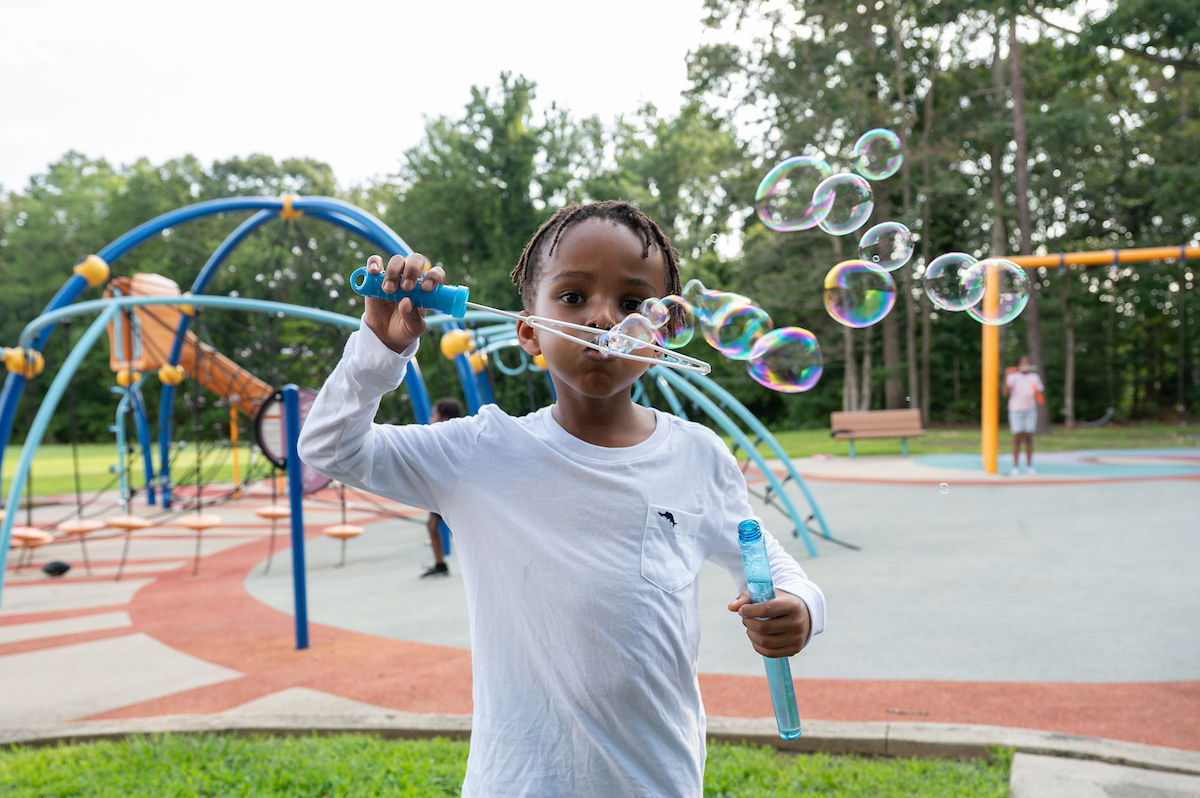 A child blows soap bubbles with a bubble wand. Behind him is a large, colorful, outdoor playground with multiple play structures and climbing elements.