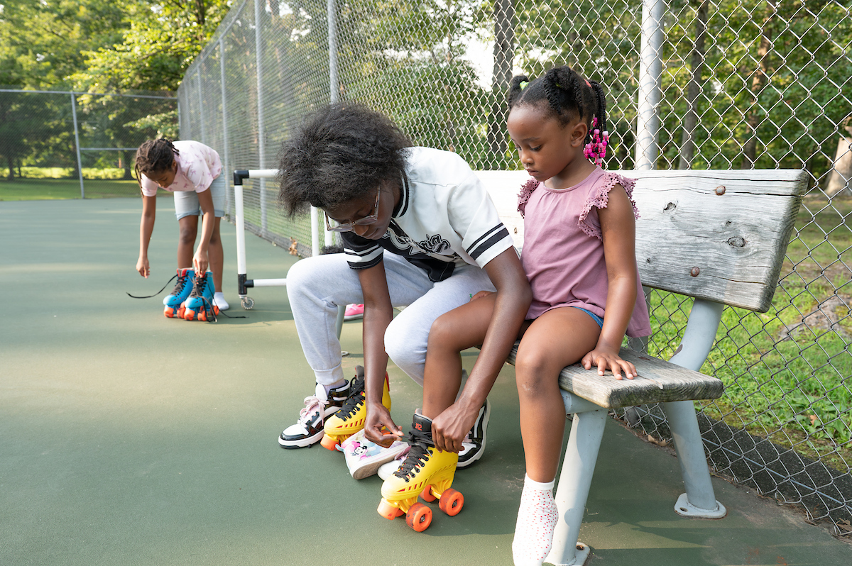 An older youth helps a child put on yellow plastic roller skates on a bench at the edge of an outdoor tennis court. Behind them, another child is putting on blue roller skates.
