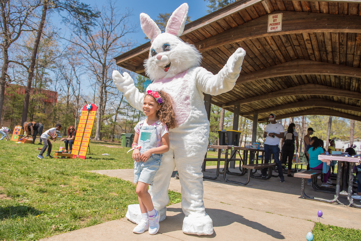 A child poses with a costumed Easter bunny in the sunshine next to a picnic shelter. Behind them in the grass, kids play carnival games.