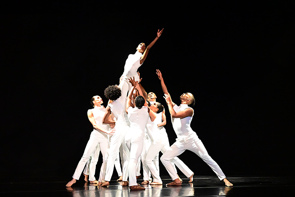 About a dozen barefoot dancers, all dressed in white pants and white sleeveless turtlenecks, dance on a black stage. Most of them surround one dancer who has her arm stretched upward, and lift her above their heads.