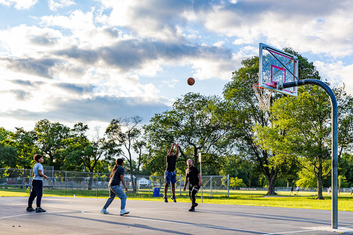 On a sunny day, four boys play basketball on an outdoor court. One boy is about a foot off the ground with his arms up, having just thrown the ball arcing toward the goal.