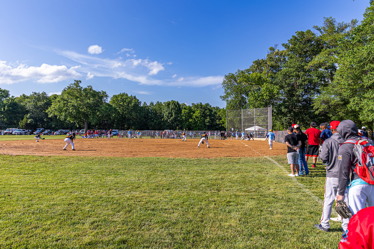 On a sunny day, a baseball game is being played on a baseball diamond in the sun. One team wears blue jerseys and the other wears black. There are trees in the distance. The photo is taken from the outfield near the third baseline.