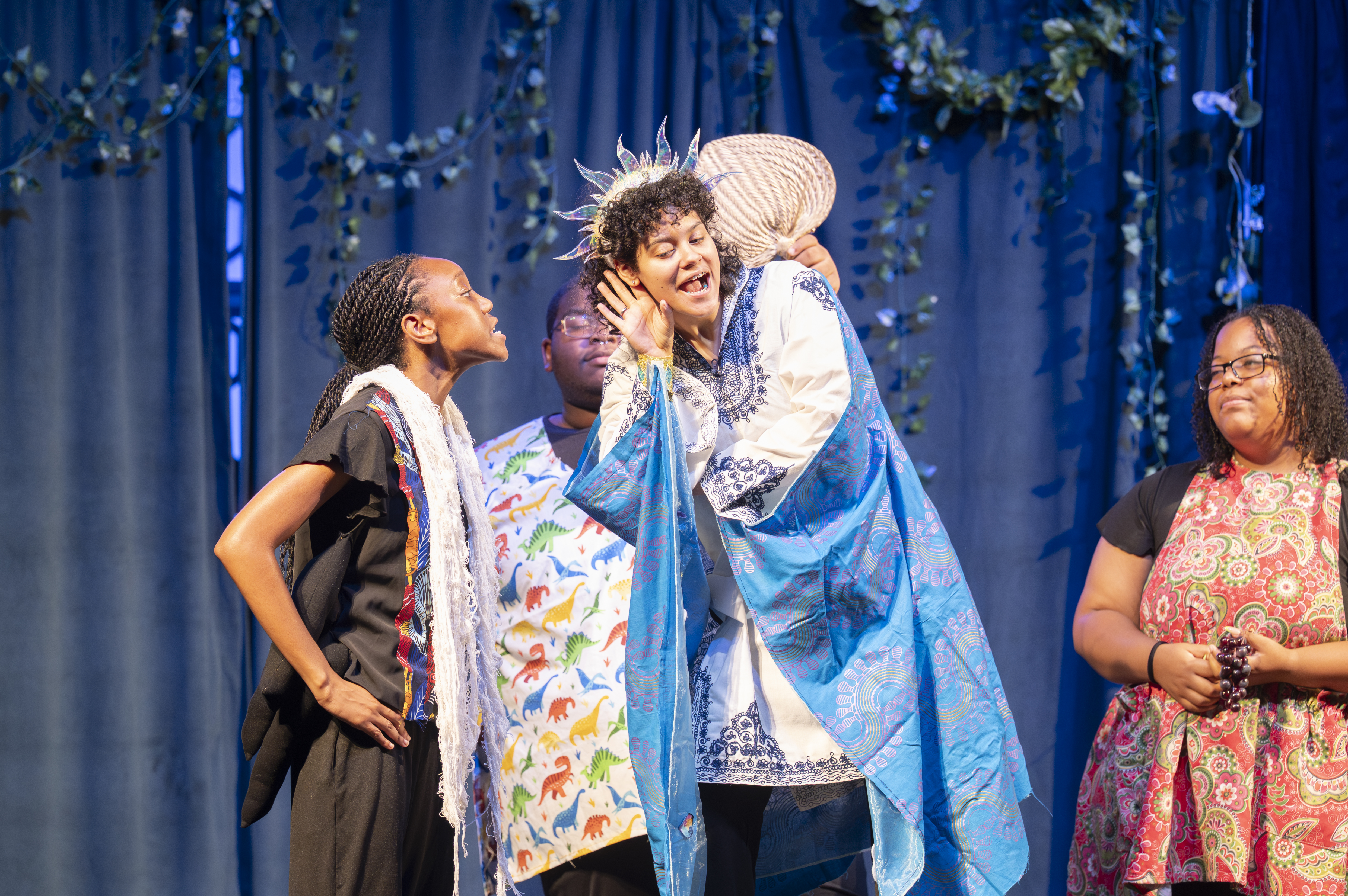 Four young actors in costumes perform on a stage in front of a dark backdrop with strands of ivy hanging from it. The central figure wears a blue cape and a spiky halo, and leans down to listen to an actor in a white scarf who is speaking.