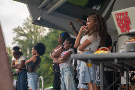 Children and adults stand on an outdoor stage during a community event, with trees and event equipment visible in the background.