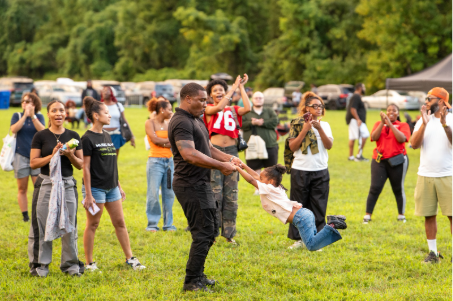 An adult spins a child by the arms on a grassy field while spectators watch and applaud nearby.