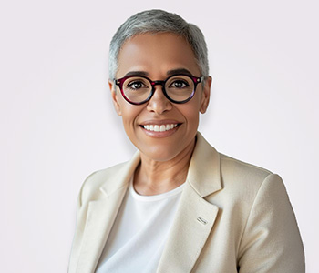 A black woman with short white hair smiling at the camera with a white background. she is wearing a white suit jacket and red glasses.