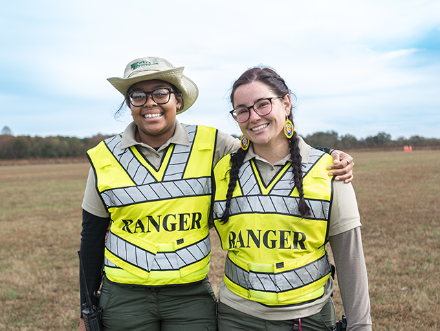 Two women standing next to each other with an arm around each other. They are both smiling and wearing high-visibility vests with the word 