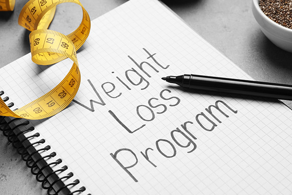A notebook on which is written the phrase "Weight Loss Program" in black marker. Next to the notebook is a tape measure and a bowl of chia seeds.