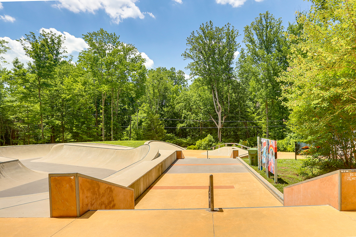 View down a short flight of stairs into a skate park with a smooth skating area, metal railings, and a large concrete bowl on the left.